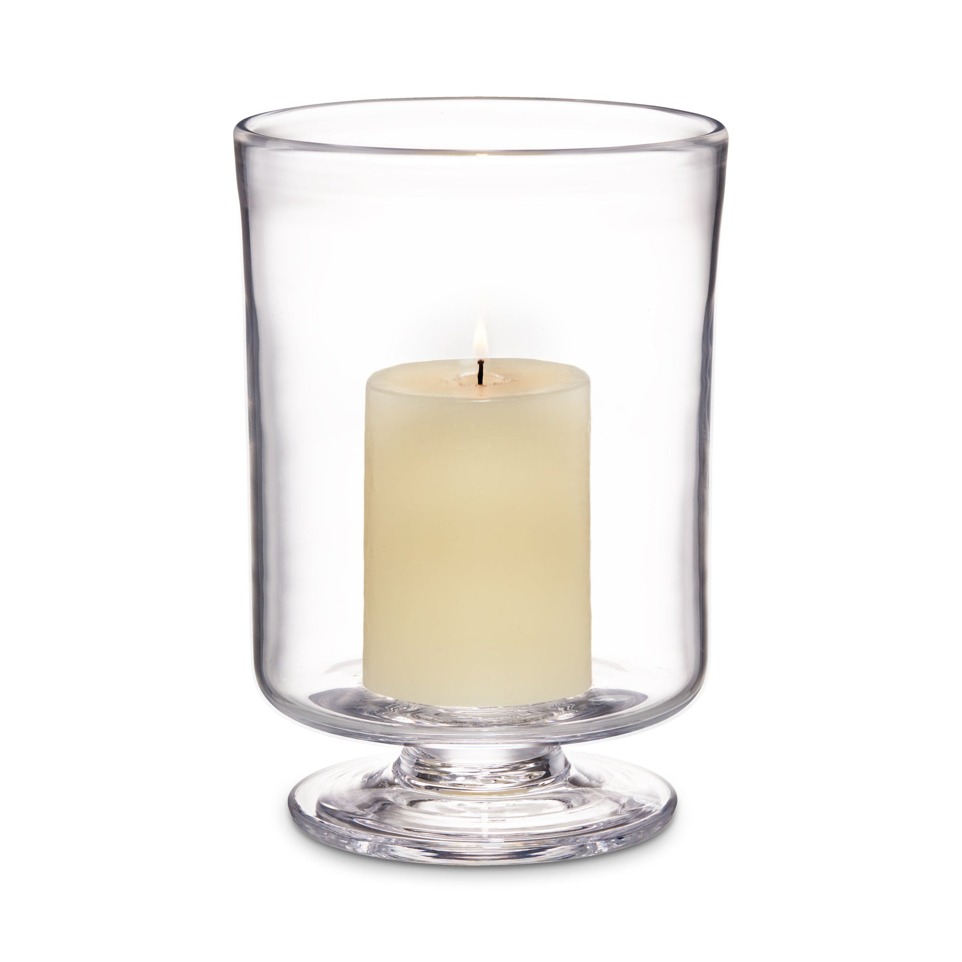 A lit cream pillar candle sits inside a clear glass hurricane holder with a pedestal base from the Nantucket Hurricane Collection by Simon Pearce, evoking the look of their classic trifle dish.