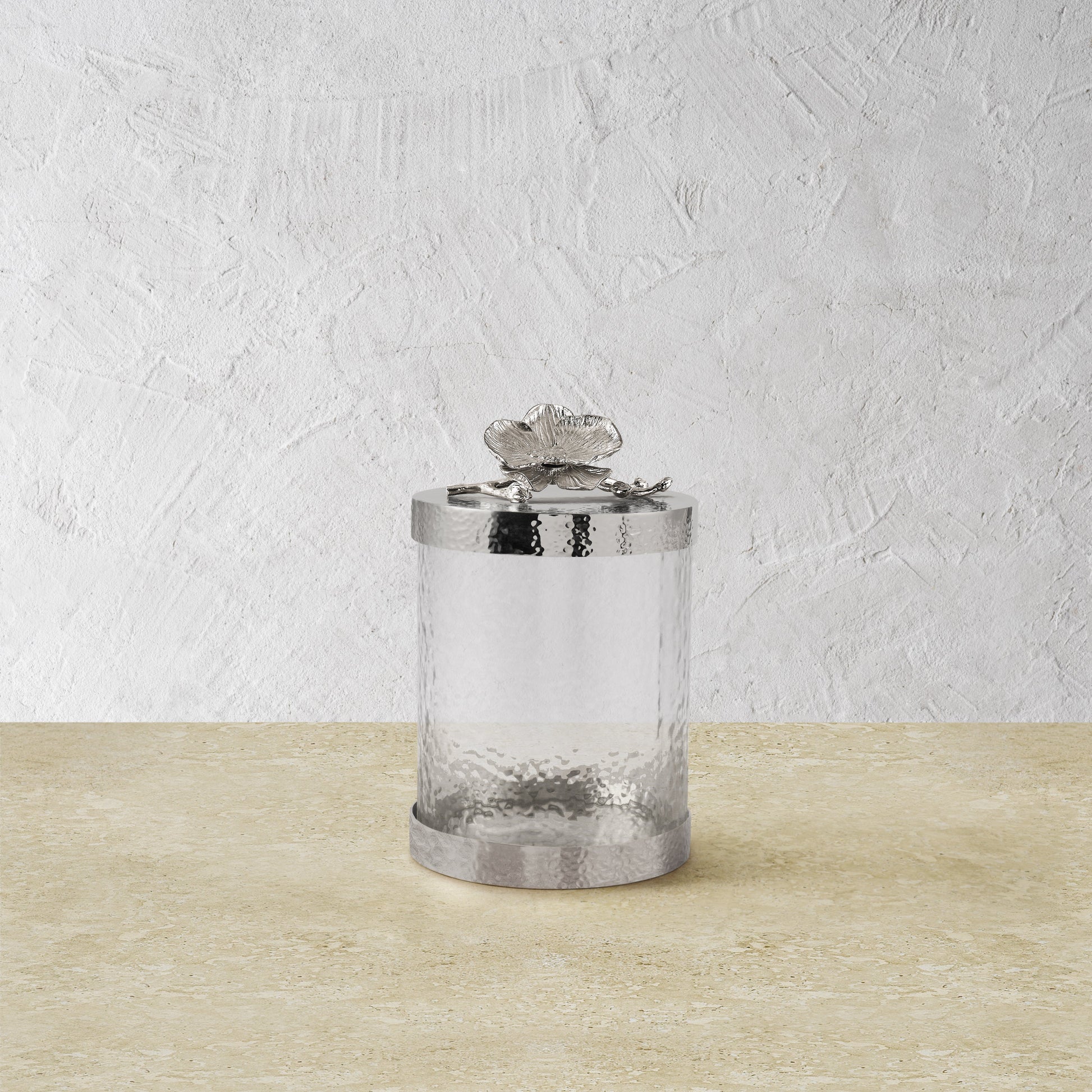 A clear, textured glass canister with a silver lid from the White Orchid Canister Collection by Michael Aram sits on a beige surface, its decorative butterfly detail reflecting refined luxury craftsmanship.