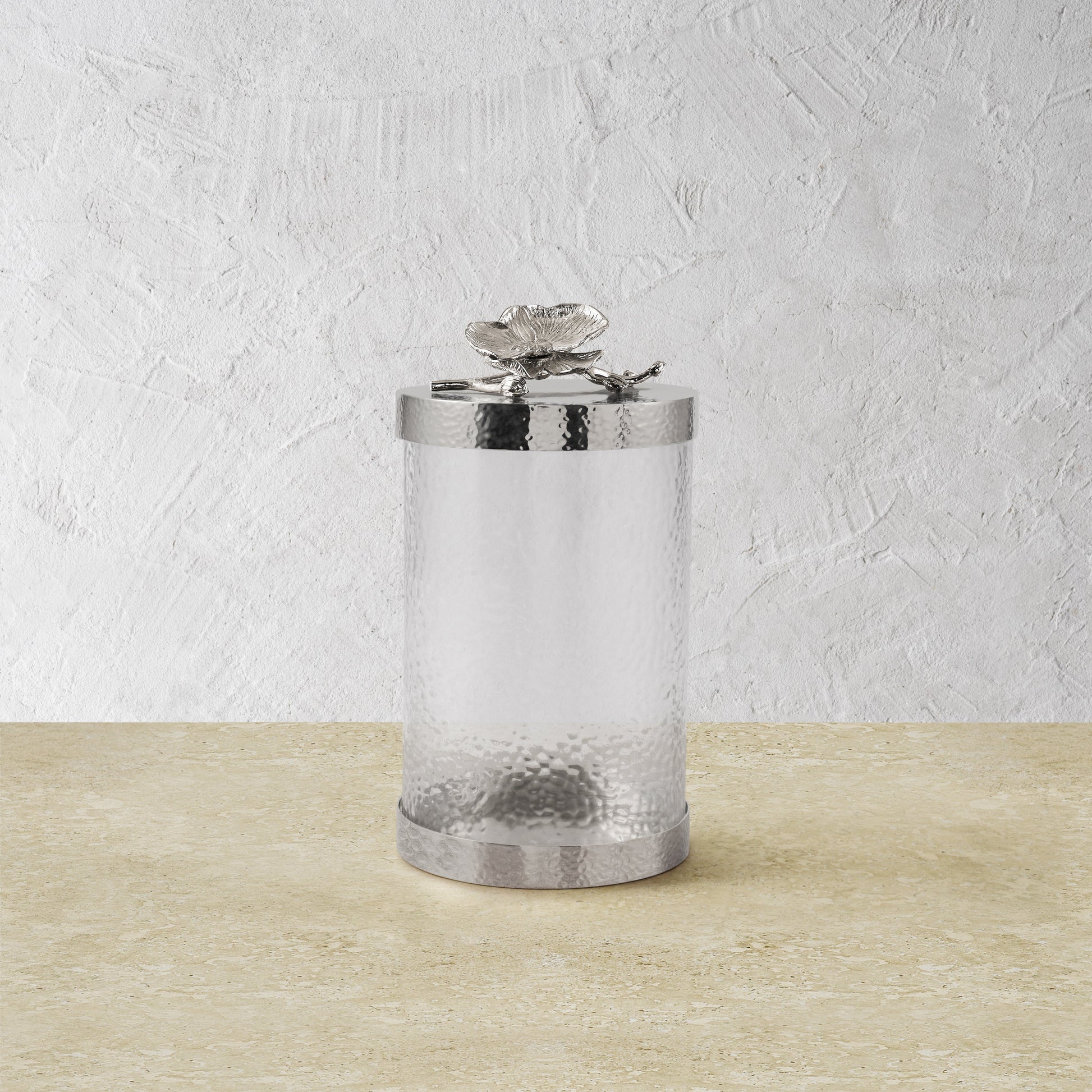 The Michael Aram White Orchid Canister Collection features a cylindrical frosted glass canister with a hammered metal lid adorned with a decorative flower, displayed on a light beige surface against a textured white wall.