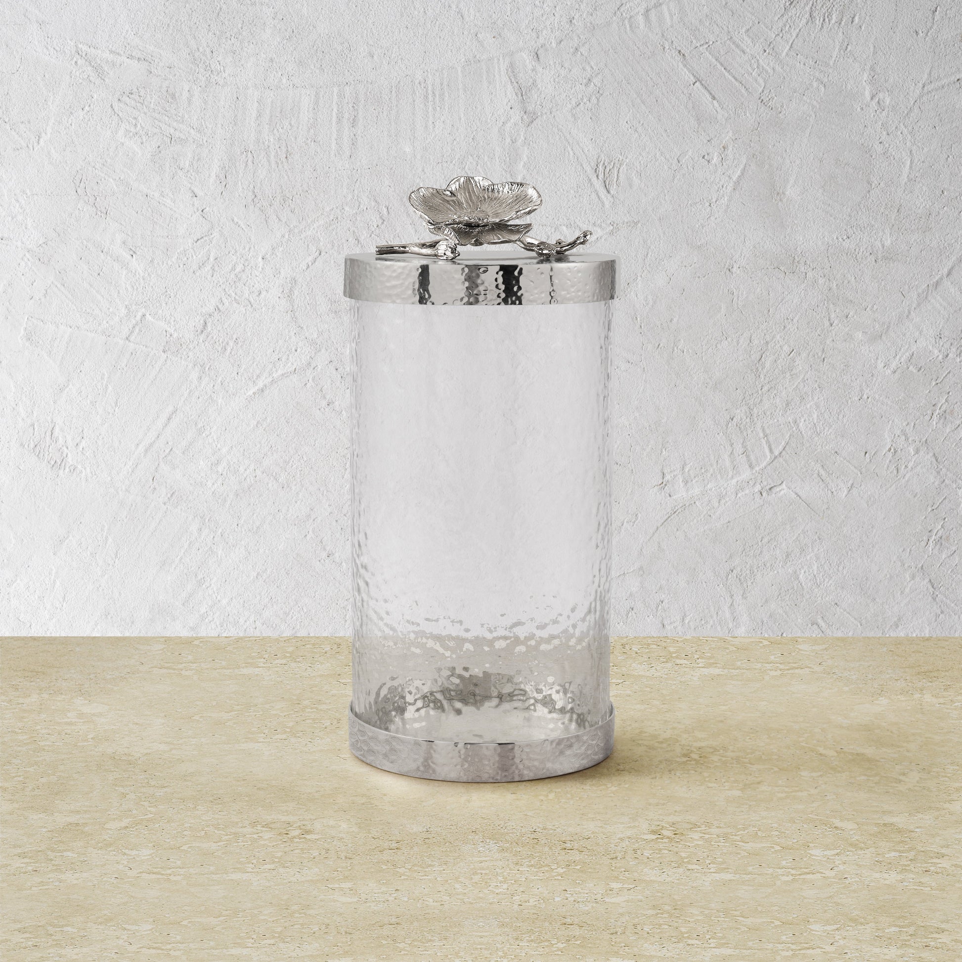The Michael Aram White Orchid Canister Collection features clear textured glass canisters with silver lids adorned with decorative flowers, showcasing luxury craftsmanship against a light beige and white textured background.