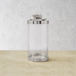 The Michael Aram White Orchid Canister Collection features clear textured glass canisters with silver lids adorned with decorative flowers, showcasing luxury craftsmanship against a light beige and white textured background.