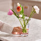 Someone places a vase from the Simon Pearce Anemone Vase Collection, filled with pink, orange, and white flowers, on a marble countertop.