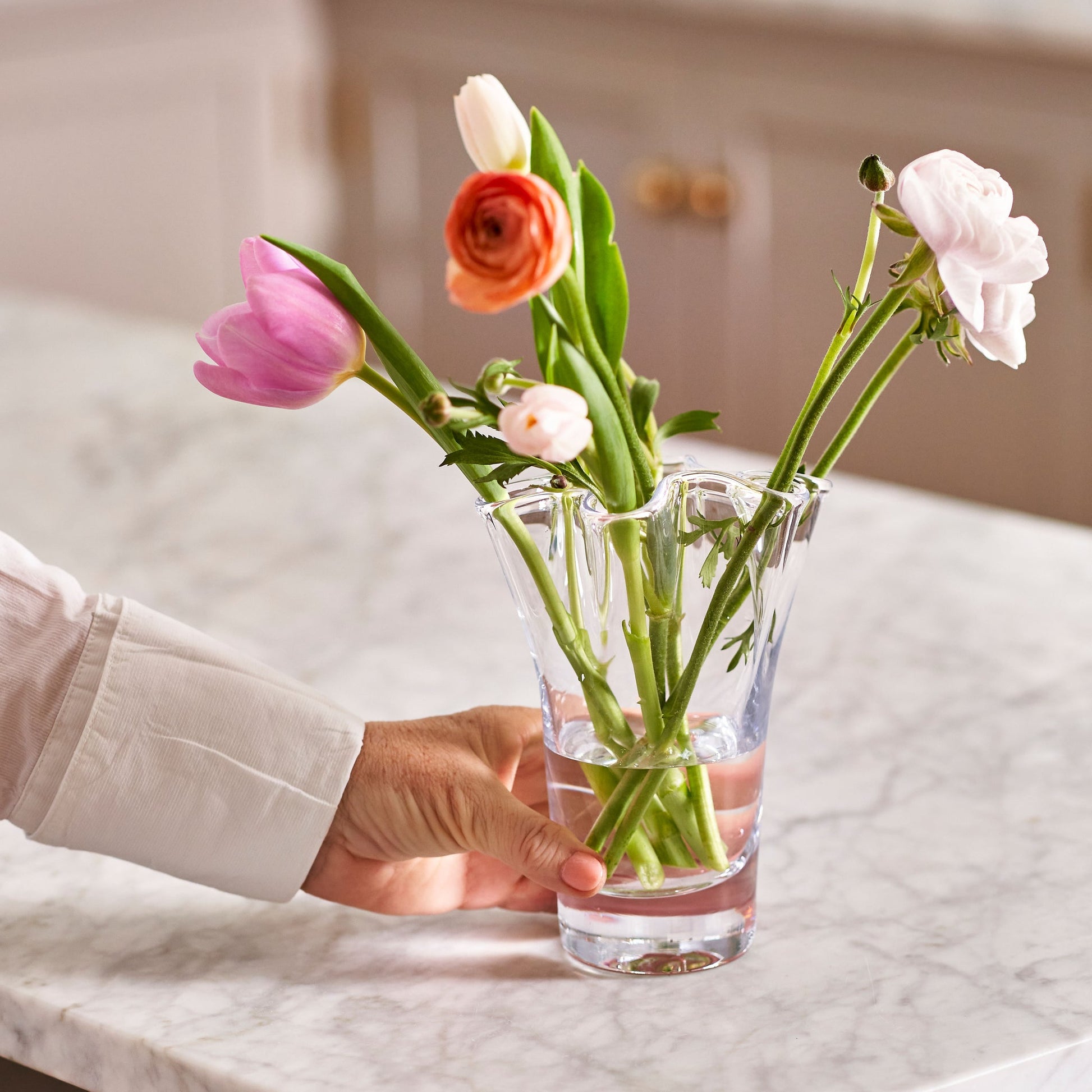 Someone places a vase from the Simon Pearce Anemone Vase Collection, filled with pink, orange, and white flowers, on a marble countertop.