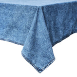 The Juliska Blue Denim Tablecloth, featuring a denim-like texture and striped edging, is draped over a rectangular table against a white background.