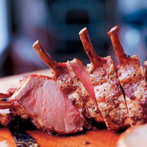 Sliced rack of cooked lamb with exposed bones, inspired by the cookbook Barefoot in Paris from Penguin Random House, resting on a wooden cutting board.