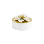 The Michael Aram Gardenia Collection features a small white round box with a gold lid adorned with a hand-enameled, white and gold gardenia jewelry flower on top.