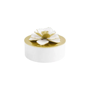 The Michael Aram Gardenia Collection features a small white round box with a gold lid adorned with a hand-enameled, white and gold gardenia jewelry flower on top.