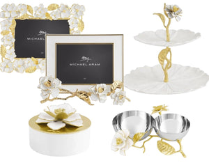 The Michael Aram Gardenia Collection features hand-enameled flowers and gold-forged leaves, with items like picture frames, a tiered tray, lidded box, and double bowl set for elegant home decor.