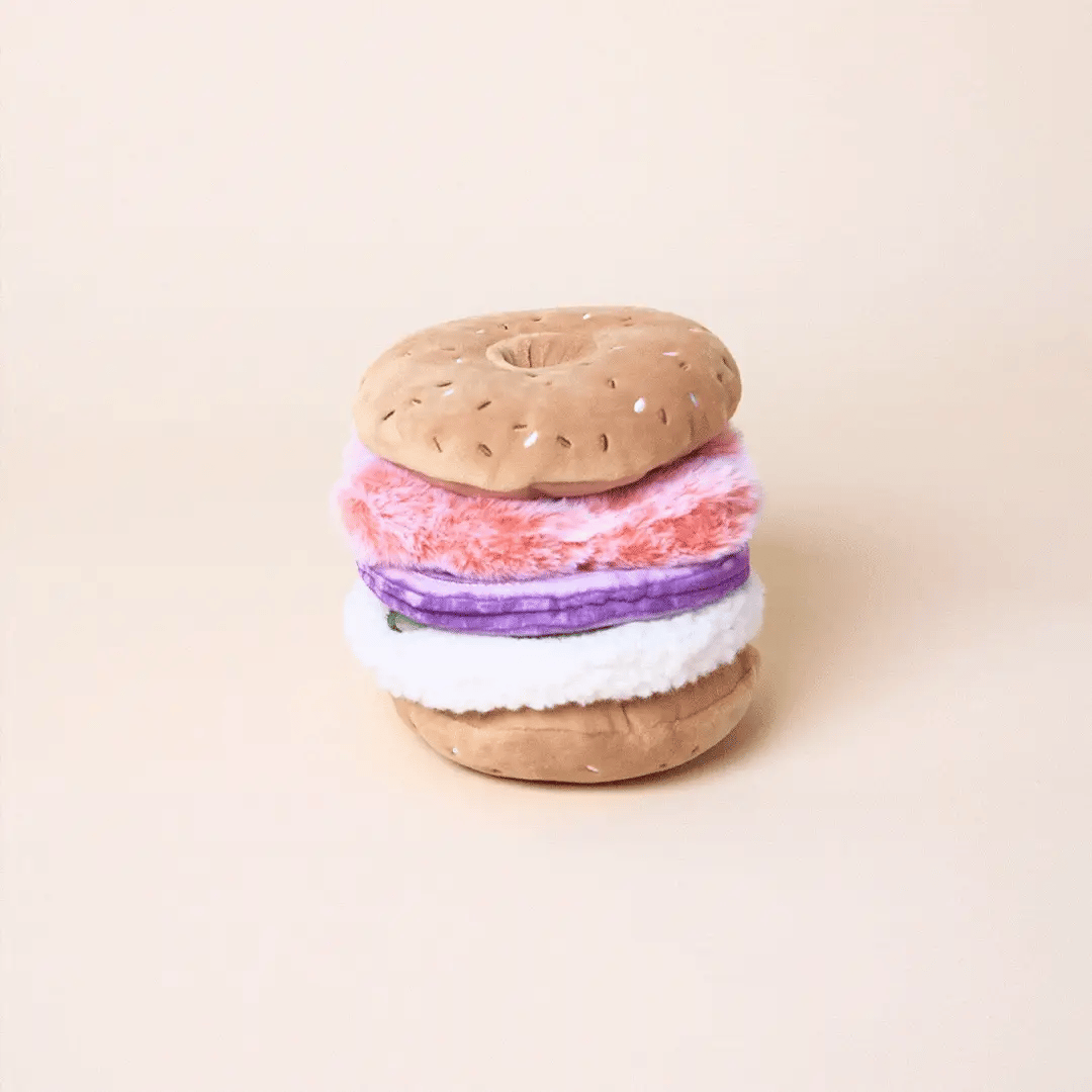 The Foggy Dog Bagel and Lox Interactive Dog Toy features pink, purple, and white layers with hidden treat pockets, crinkle, and squeak elements for added fun, shown on a plain beige background.