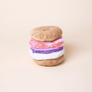 The Foggy Dog Bagel and Lox Interactive Dog Toy features pink, purple, and white layers with hidden treat pockets, crinkle, and squeak elements for added fun, shown on a plain beige background.