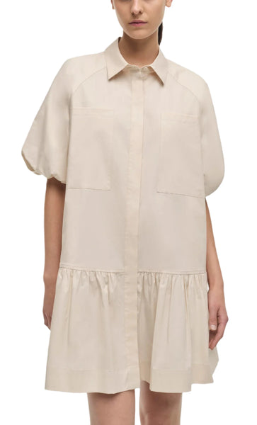 A person wearing a Simkhai Crissy Dress with a loose-fitting, beige design featuring ballooned short sleeves, a collar, and two front pockets. The mini dress has a gathered section above the hemline for added flair.