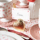 A place card reading "Siena" sits in Juliska's Berry & Thread Heart Napkin Ring and Place Card Holder, atop a table with pink dishware, hammered accents, and heart-patterned decor.