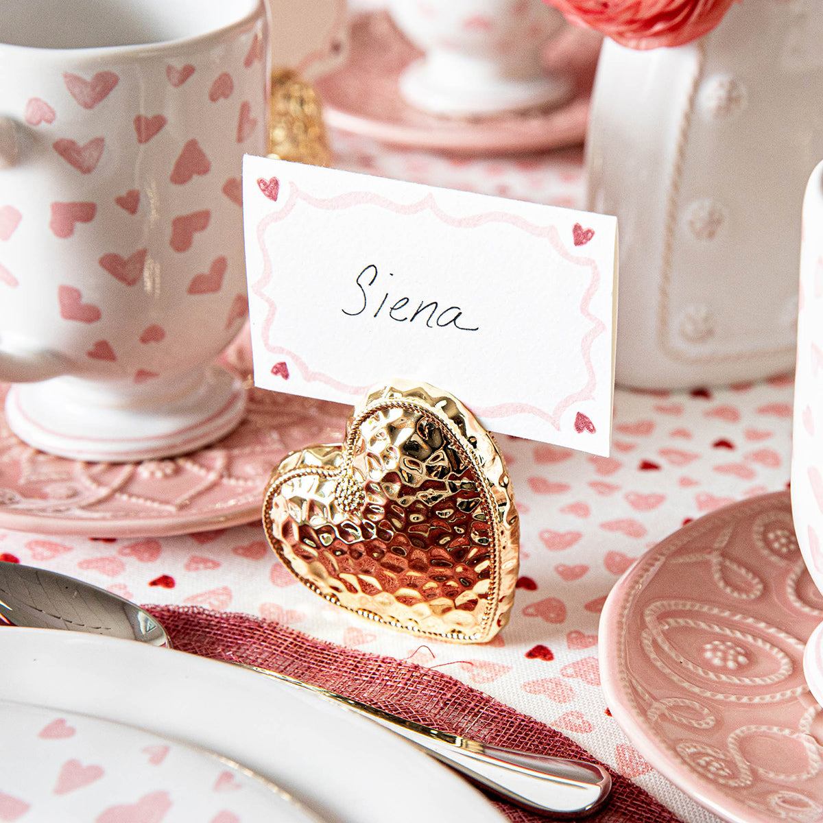 A place card reading "Siena" sits in Juliska's Berry & Thread Heart Napkin Ring and Place Card Holder, atop a table with pink dishware, hammered accents, and heart-patterned decor.