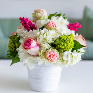 The Live Mother's Day Floral Arrangement by Hive Floral Studio features vibrant white, pink, and green roses, carnations, and hydrangeas in a white ceramic vase, beautifully displayed on a white table with a blurred sofa in the background.