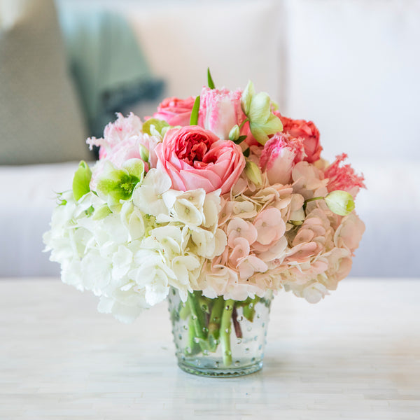 The Live Valentine's Floral Arrangement by HIVE Home, Gift and Garden features pink roses, white hydrangeas, and light green seasonal blooms in a glass vase on a white surface against a blurred background.