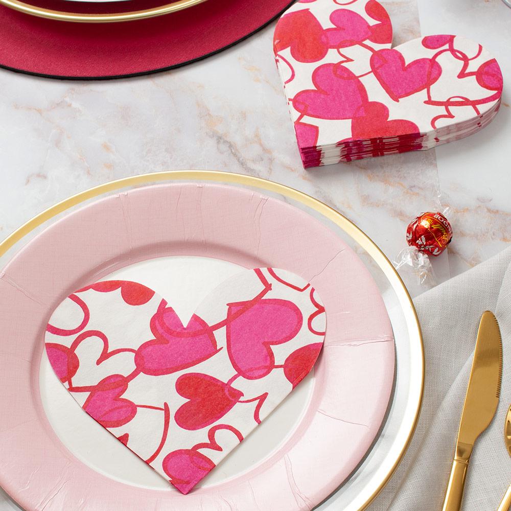 A pink plate with a gold rim holds a heart-shaped Caspari Painted Hearts Luncheon Napkin; more of these paper linen napkins and a gold utensil are nearby.