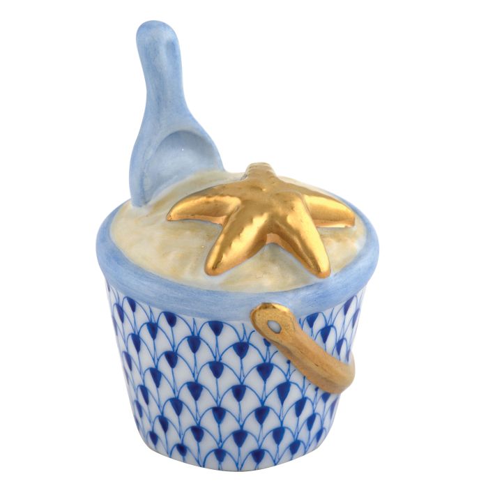 The Herend Beach Bucket Collection features a hand-painted white porcelain bucket with blue patterns, filled with white material, topped with a gold starfish, and includes a blue scoop inside.
