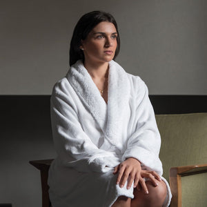 A woman wearing the Graccioza Egoist Bath Robe, made from combed cotton, sits on a chair with her hands on her knee, gazing ahead with a neutral expression.