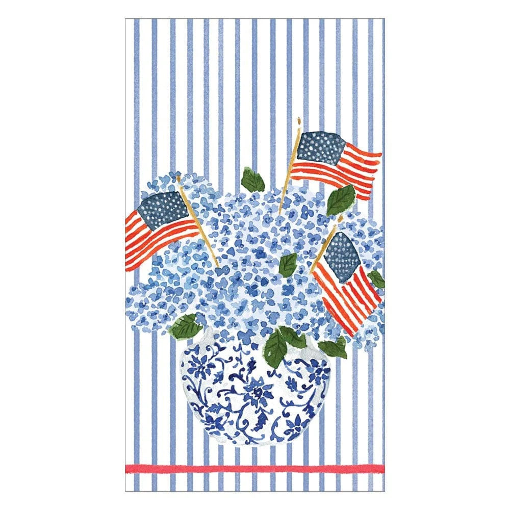 The Flags And Hydrangeas Collection by Caspari features blue hydrangeas and American flags in a floral vase, set on a picnic table with blue, white, and red stripes—perfect for summer.