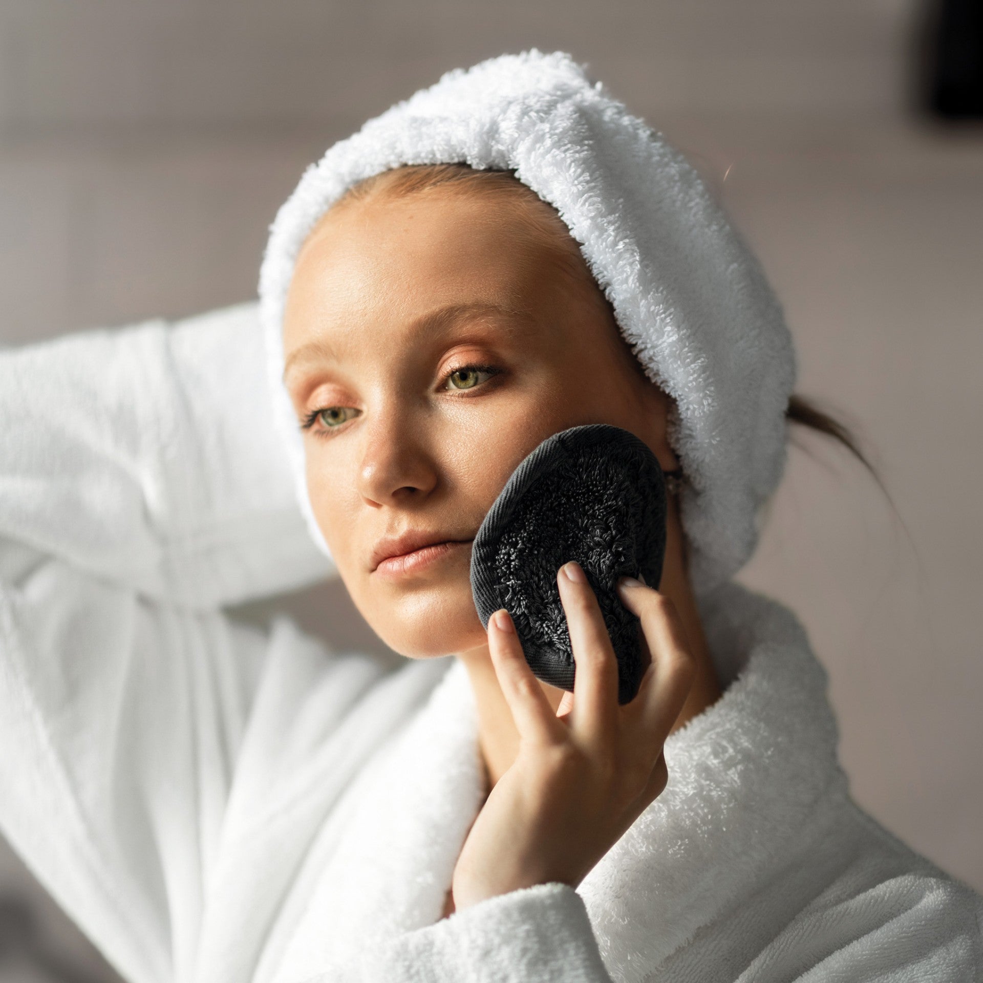 Wearing a GRACCIOZA Egoist Headband in white, a person uses a black textured facial cleansing pad on their cheek.
