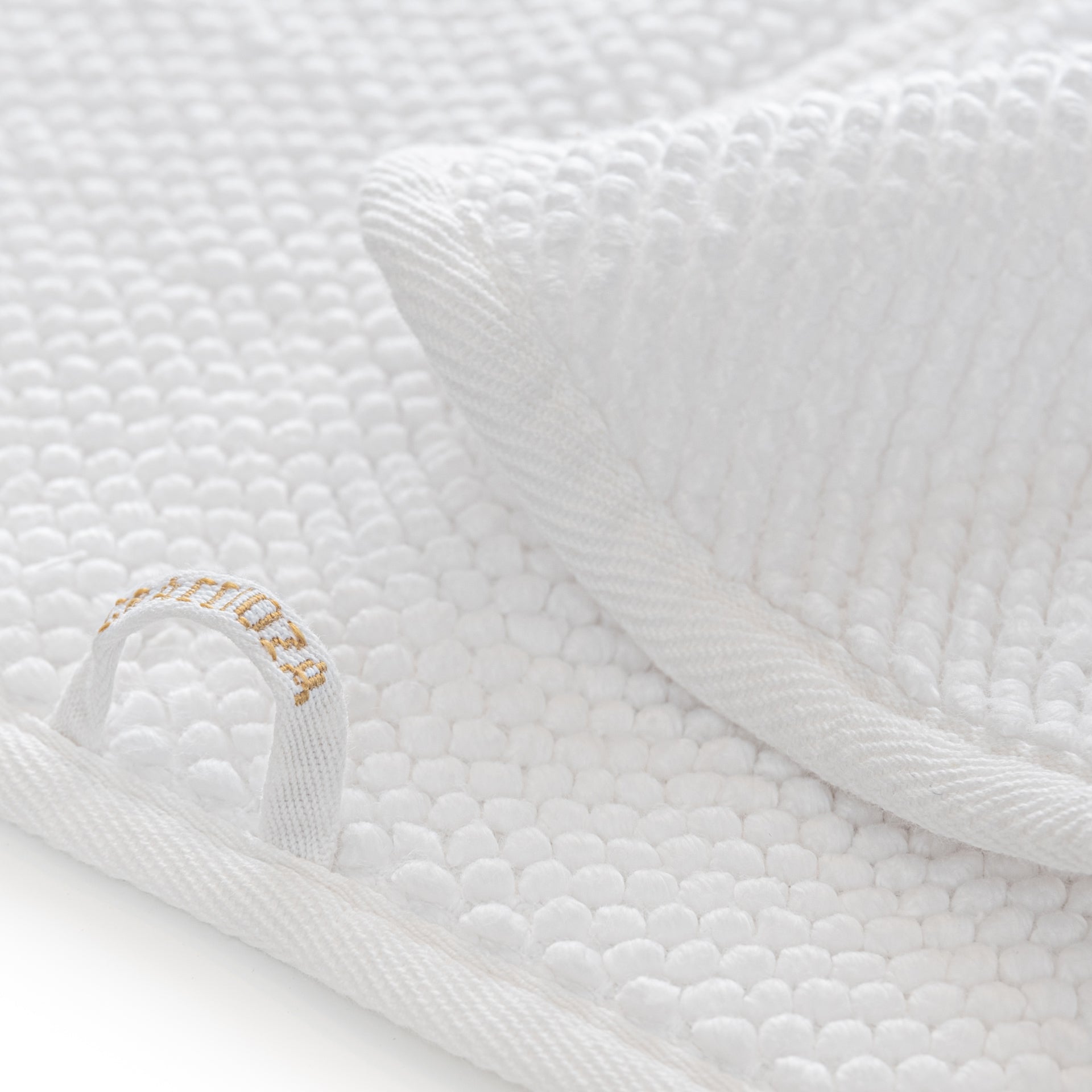 Close-up of an ultra-soft, white waffle-textured towel from the Graccioza Pearls Bath Collection by Gracciozza, featuring a decorative loop on one corner and embroidered with gold text.