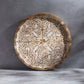 The Mya Inlay Serving and Decor Collection by Be Home features a round tray with an acacia wood rim and gold-beige leaf pattern on white, displayed on a gray textured surface.