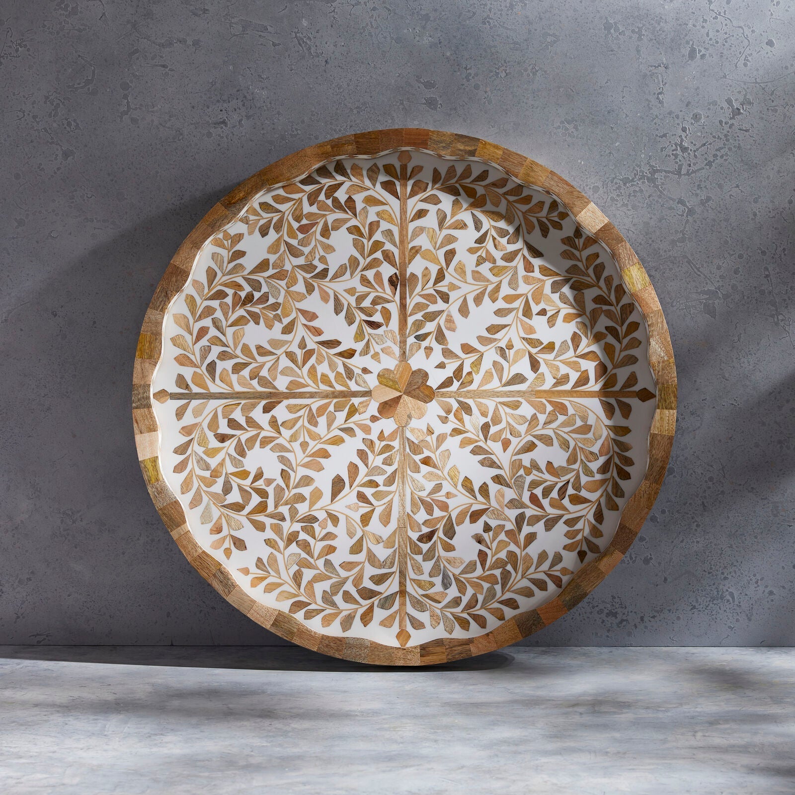 The Mya Inlay Serving and Decor Collection by Be Home features a round tray with an acacia wood rim and gold-beige leaf pattern on white, displayed on a gray textured surface.