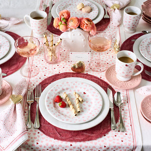 A table set with Juliska's Love You More Decor and Tabletop Collection features pink heart plates, pink placemats, glasses of rosé, wafer cookies, strawberries, and floral accents in a romantic pink and white theme.