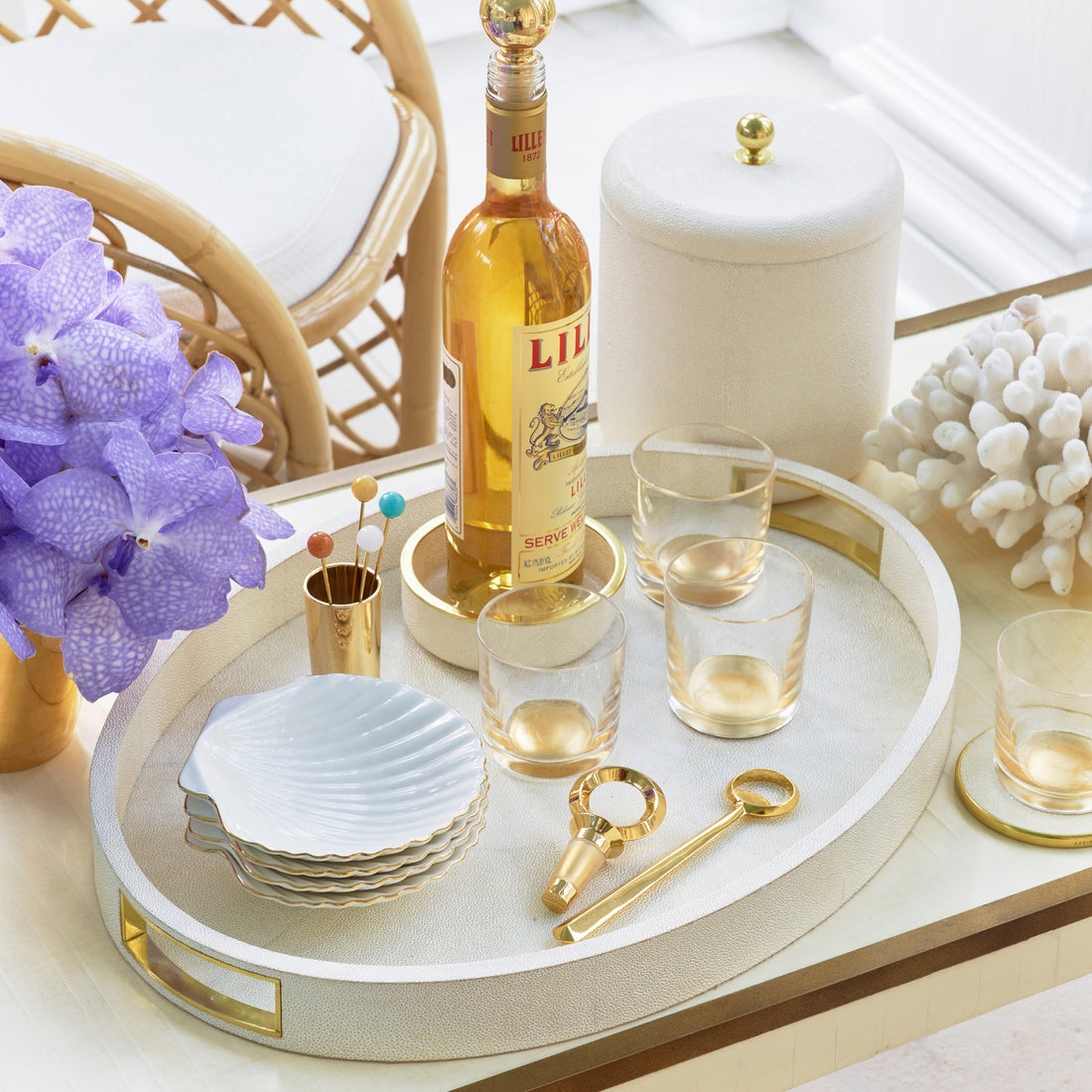 A tray on the table displays a bottle of Lillet, four gold-edged glasses, a gold corkscrew, cocktail picks, a cup, and an Aerin Shell Appetizer Plate, with a vase of purple flowers and coastal coral decor nearby.