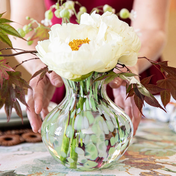 A person arranges white peonies and greenery in the Juliska Puro Speckled Vase, displaying the brand's glass artistry on a patterned tablecloth.