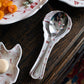 A metal slotted serving spoon rests on the Juliska Country Estate Winter Frolic Ruby Spoon Rest, featuring festive red patterns, set on a dark wooden table—an ideal touch for holiday gatherings.