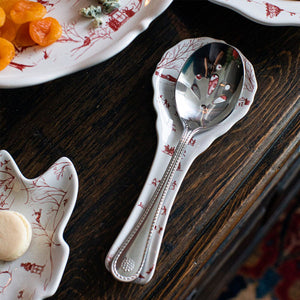 A metal slotted serving spoon rests on the Juliska Country Estate Winter Frolic Ruby Spoon Rest, featuring festive red patterns, set on a dark wooden table—an ideal touch for holiday gatherings.