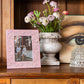An Addison Ross Chinoiserie Frame in pink is artfully arranged on a wooden shelf beside a vase of purple and white flowers, Sarah David Ross's book with an eye cover, and a textured high-gloss lacquer object.
