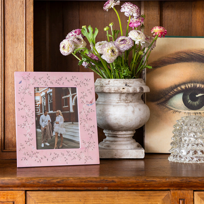An Addison Ross Chinoiserie Frame in pink is artfully arranged on a wooden shelf beside a vase of purple and white flowers, Sarah David Ross's book with an eye cover, and a textured high-gloss lacquer object.