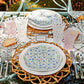 A table set for a meal features floral plates, woven placemats, cake slices, and Juliska's Trellis Drinkware Collection with pink and clear lemonade glasses on a floral tablecloth, plus a name card reading "Olivia.