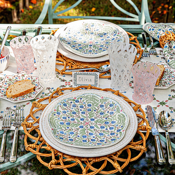 A table set for a meal features floral plates, woven placemats, cake slices, and Juliska's Trellis Drinkware Collection with pink and clear lemonade glasses on a floral tablecloth, plus a name card reading "Olivia.