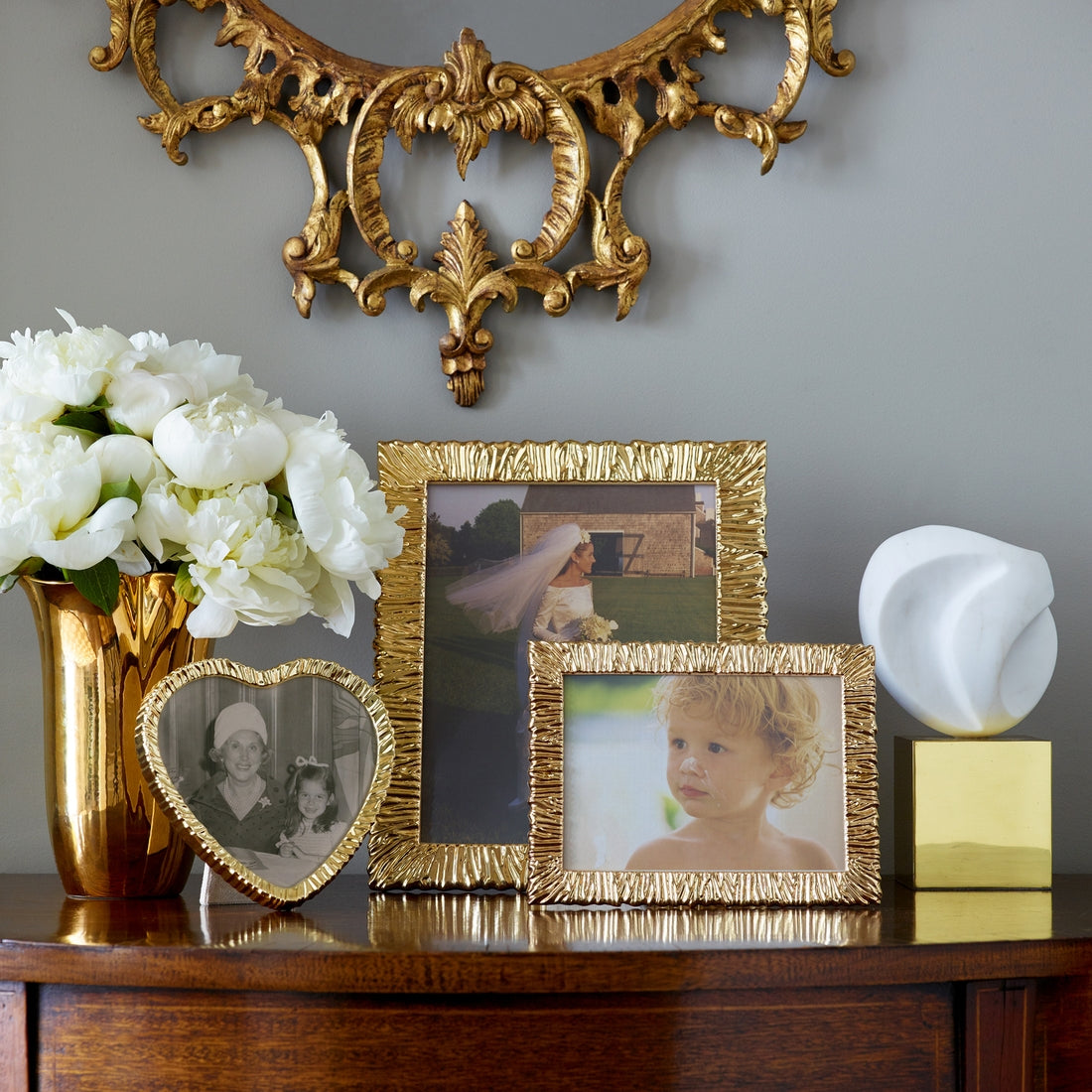 A wooden table holds a gold vase with white flowers, three Aerin Ambroise Frame Collection photo frames, a white abstract sculpture, and a textured gold-framed mirror mounted on the wall above.