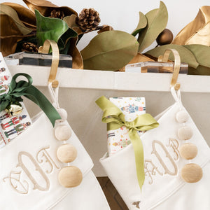 Two white stockings adorned with initials are suspended from a mantle using Fig and Dove Nickel & Acrylic Stocking Holders. Each stocking is filled with wrapped gifts tied with bows. The mantle above is decorated with pinecones and greenery.