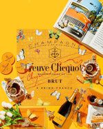 A completed Acrylic Puzzle by Tart by Taylor is surrounded by a champagne bottle, teacups, tulips, a cigar, orange accessories, and an open magazine on a bright yellow surface—perfect for stylish home decor.