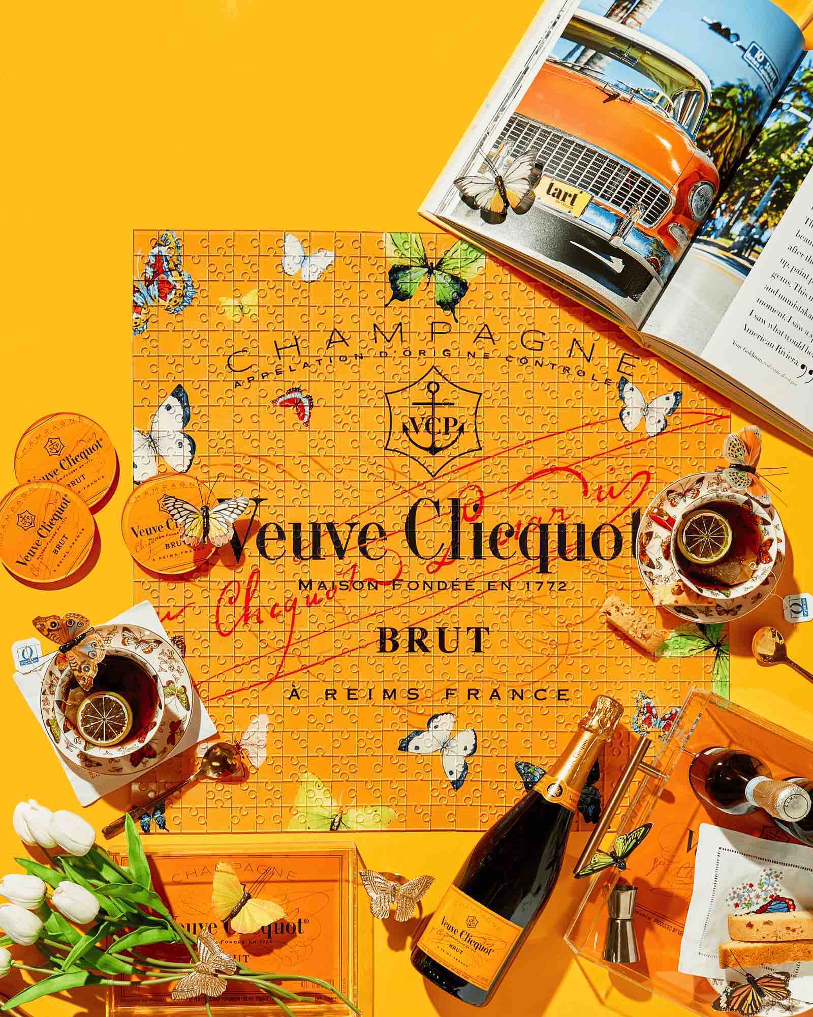 A completed Acrylic Puzzle by Tart by Taylor is surrounded by a champagne bottle, teacups, tulips, a cigar, orange accessories, and an open magazine on a bright yellow surface—perfect for stylish home decor.
