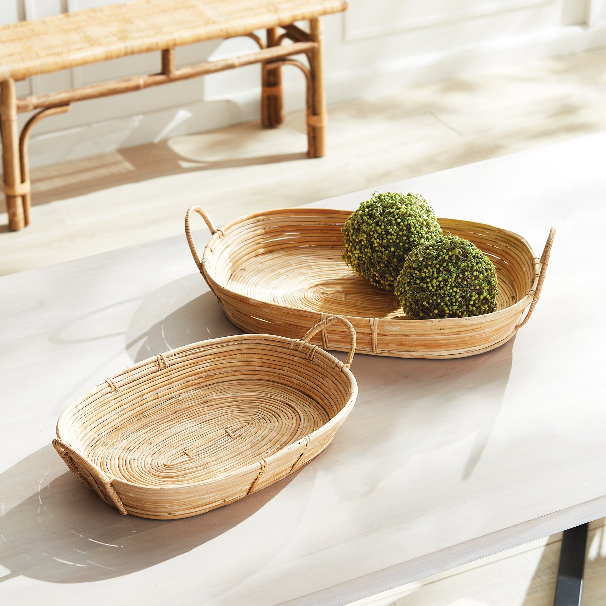 Two trays from the Cane Rattan Tray Collection by Napa Home & Garden sit on a white table; one holds two artificial green decorative spheres, while the other is empty.