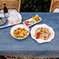 A Juliska Blue Denim Tablecloth is set outdoors, topped with grilled chicken, salad, vegetables, glasses, plates, and a bottle of wine on a tray.