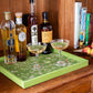 The Medium Chinoiserie Tray Collection by Addison Ross, shown in high gloss lacquer on wood, displays two glasses with pale yellow drinks, four liquor bottles, a peach, and several books on a shelf in the background.