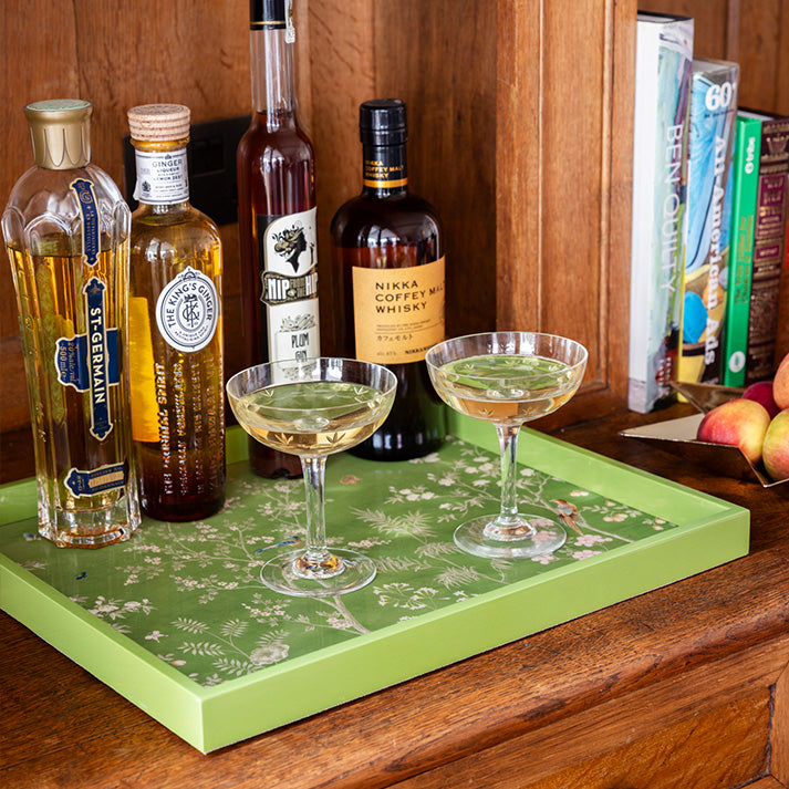 The Medium Chinoiserie Tray Collection by Addison Ross, shown in high gloss lacquer on wood, displays two glasses with pale yellow drinks, four liquor bottles, a peach, and several books on a shelf in the background.