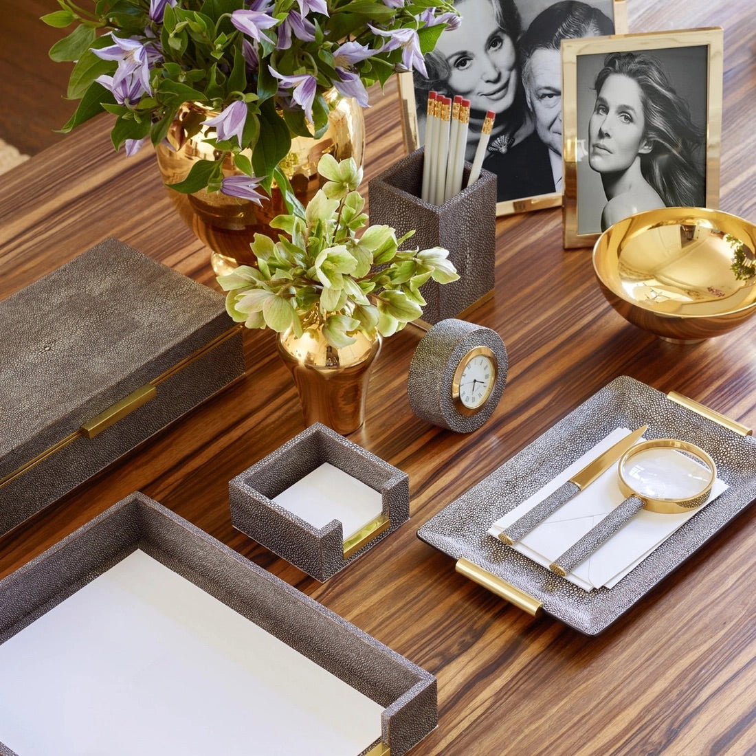 A wooden desk organized with an Aerin Shagreen Small Vanity Tray in Chocolate, gold vases with flowers, a clock, brass handles, framed photos, a pen holder with pens, and a magnifying glass on a napkin.