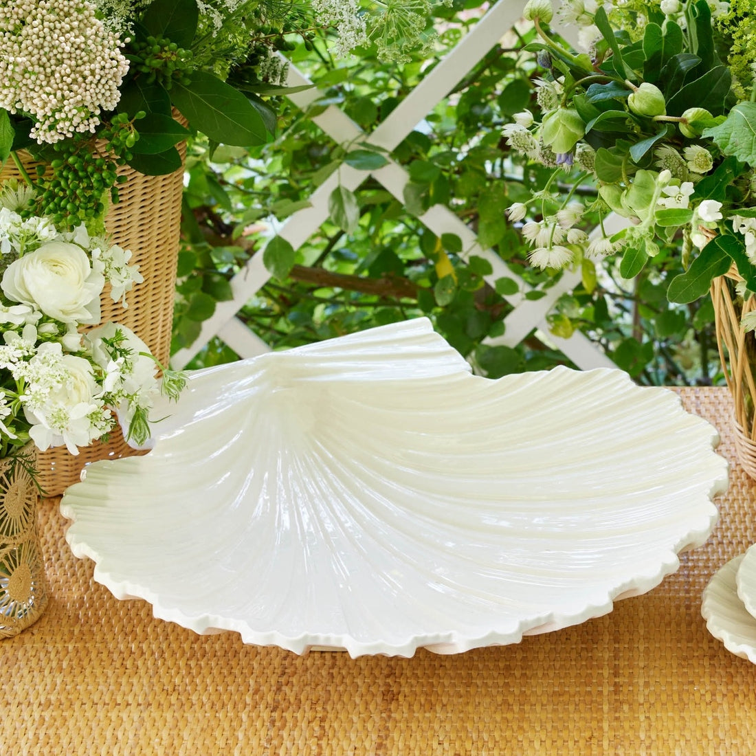 The Cream Shell Platter Collection by Aerin features a glossy white earthenware serving platter, showcased on a woven surface with baskets and white flowers—an ideal accent for coastal-inspired decor.
