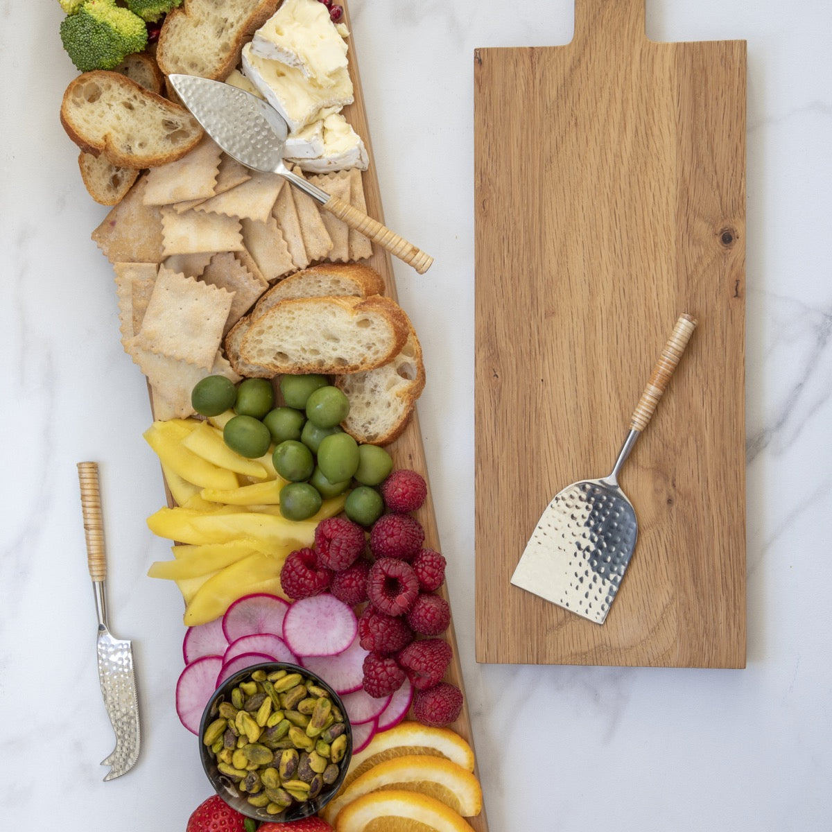 The Be Home Aquila Serving Collection features a wooden board topped with bread, crackers, cheese, fruits, raspberries, olives, radishes, pistachios, and quality cutlery beside an empty cutting board on a white surface.