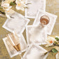 The Kim Seybert Pin Dot Cocktail Napkin Set, featuring six white elegant napkins with decorative stitching, is displayed on a gold surface alongside two glasses and white magnolia flowers for a sophisticated touch.