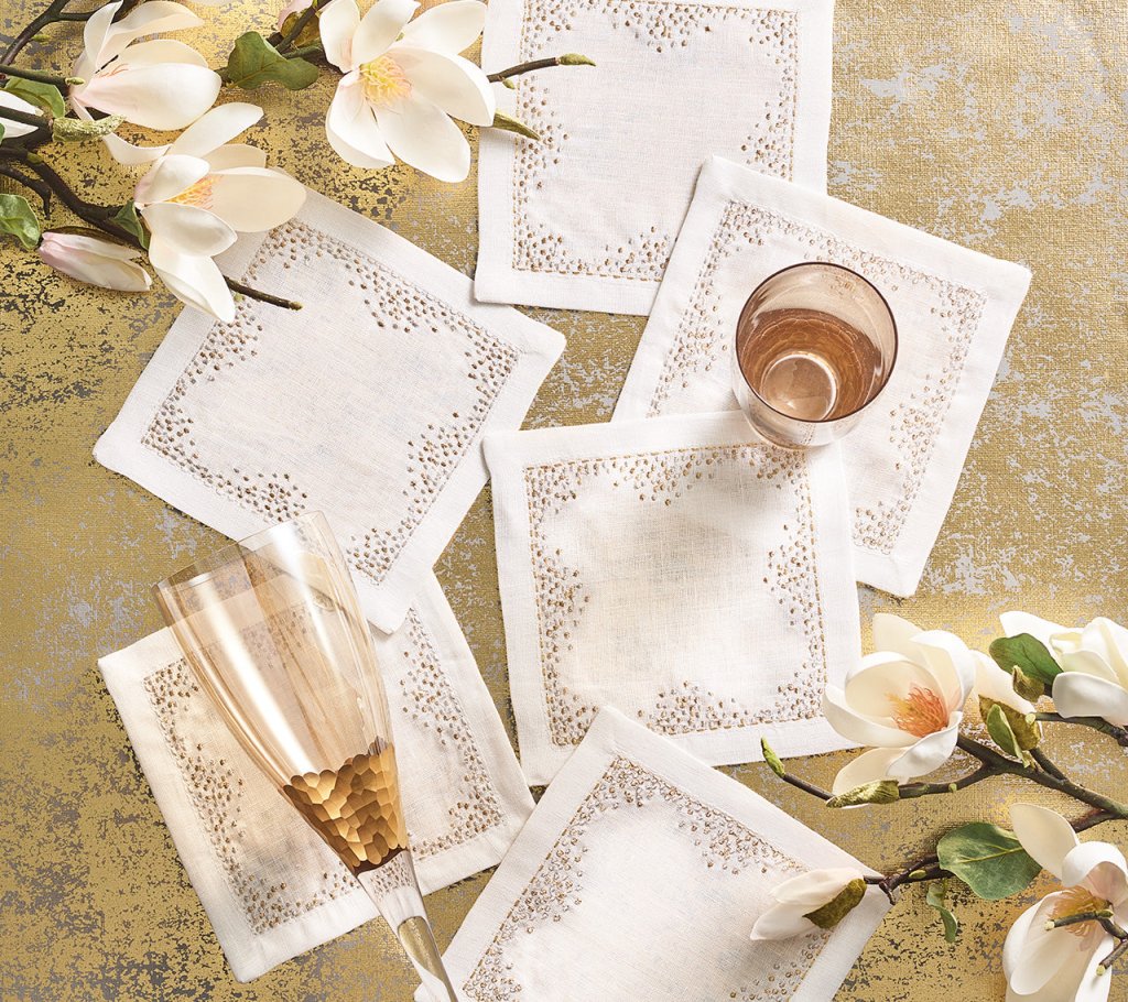 The Kim Seybert Pin Dot Cocktail Napkin Set, featuring six white elegant napkins with decorative stitching, is displayed on a gold surface alongside two glasses and white magnolia flowers for a sophisticated touch.