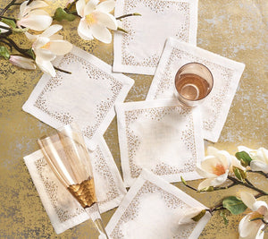 The Kim Seybert Pin Dot Cocktail Napkin Set, featuring six white elegant napkins with decorative stitching, is displayed on a gold surface alongside two glasses and white magnolia flowers for a sophisticated touch.